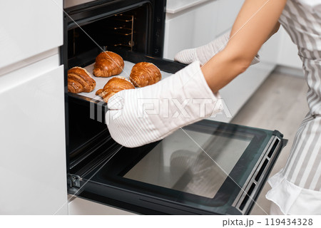 female baker taking tray with fresh croissants from oven in modern kitchen, close-up 119434328