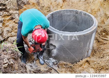Using rotary hammer with chisel, worker punches hole for sewer pipe in concrete septic tank ring. 119435997