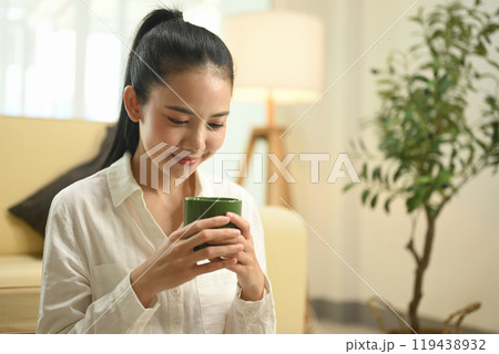 Happy young woman sitting in living room, holding a warm green mug with both hands and smiling softly 119438932