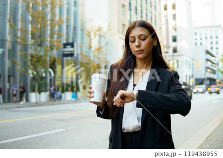 Beautiful young businesswoman holding coffee cup and checking time on her watch while walking down an urban street Beautiful young businesswoman holding coffee cup and checking time on her watch while walking down an urban street 119438955