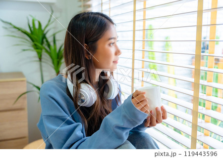 Young asian woman standing beside window and holding mug in bedroom at home Young asian woman standing beside window and holding mug in bedroom at home 119443596