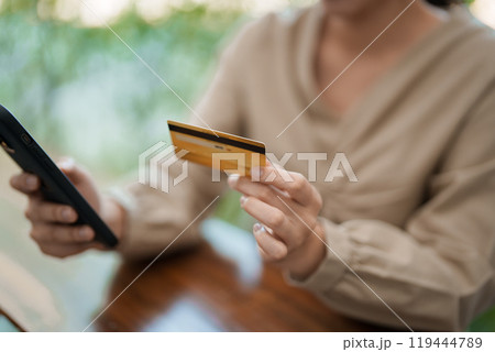 A young woman is sitting at her desk, smiling as she holds her phone and a credit card, likely making an online purchase. She is in a modern, bright office environment A young woman is sitting at her desk, smiling as she holds her phone and a credit card, likely making an online purchase. She is in a modern, bright office environment 119444789