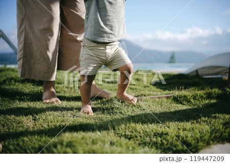 A toddler is taking their first steps on soft, green grass with adult support 119447209