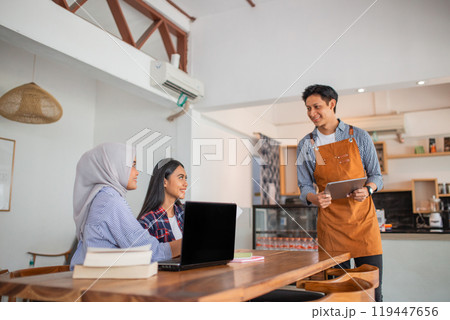 two women work on laptops while ordering food from a male waiter 119447656