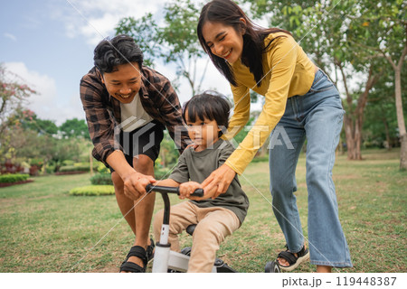 little boy learning to ride a bike with mom and dad outdoors 119448387