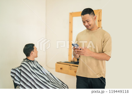 A man gets his haircut at a barber shop while another checks his phone 119449118