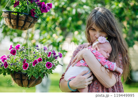 Young mother gently cradles her sleeping newborn baby girl amidst lovely hanging flower baskets in a lush garden 119449995