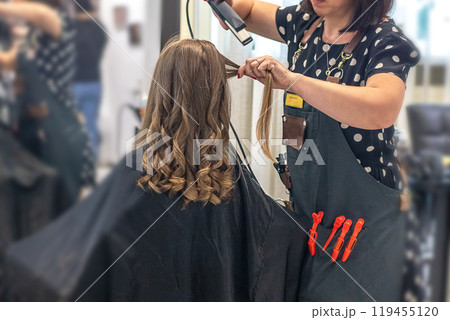 Hands of a hairdresser with a curling iron doing a girl's hair in a professional beauty salon 119455120