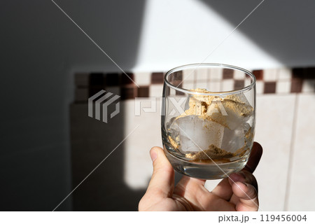A glass filled with ice cubes and powdered Amanita muscaria mushroom on its surface, held in the hand, illuminated by natural sunlight. Preparing a homemade fly agaric elixir A glass filled with ice cubes and powdered Amanita muscaria mushroom on its surface, held in the hand, illuminated by natural sunlight. Preparing a homemade fly agaric elixir 119456004