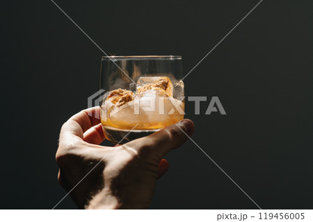 A glass filled with ice cubes and powdered Amanita muscaria mushroom on its surface, held in the hand, illuminated by natural sunlight. Preparing a homemade fly agaric elixir 119456005