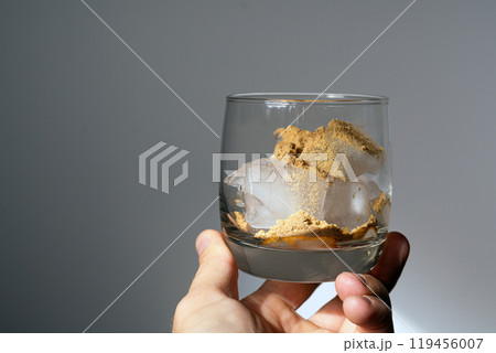 A glass filled with ice cubes and powdered Amanita muscaria mushroom on its surface, held in the hand, illuminated by natural sunlight. Preparing a homemade fly agaric elixir A glass filled with ice cubes and powdered Amanita muscaria mushroom on its surface, held in the hand, illuminated by natural sunlight. Preparing a homemade fly agaric elixir 119456007