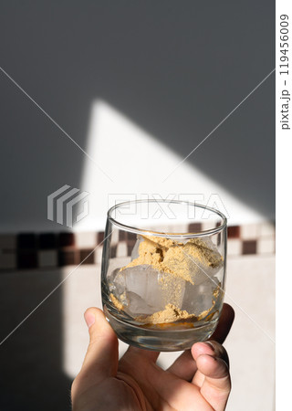 A glass filled with ice cubes and powdered Amanita muscaria mushroom on its surface, held in the hand, illuminated by natural sunlight. Preparing a homemade fly agaric elixir 119456009
