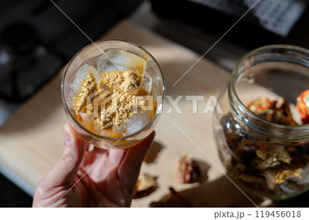 A glass filled with ice cubes and powdered Amanita muscaria mushroom on its surface, held in the hand, illuminated by natural sunlight. Preparing a homemade fly agaric elixir 119456018