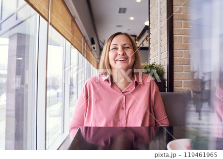Cheerful woman in pink shirt smiling at cafe table by large window on a bright Cheerful woman in pink shirt smiling at cafe table by large window on a bright 119461965
