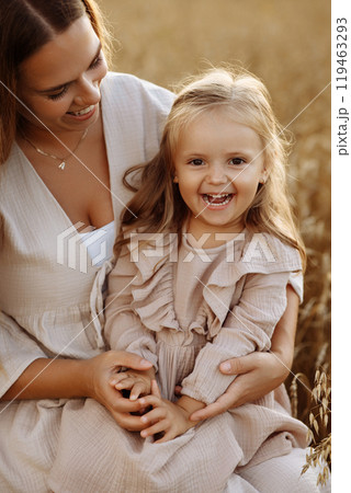 A little blonde girl in a beige dress hugs her mother in the rays of a warm summer sunset in a wheat field. 119463293