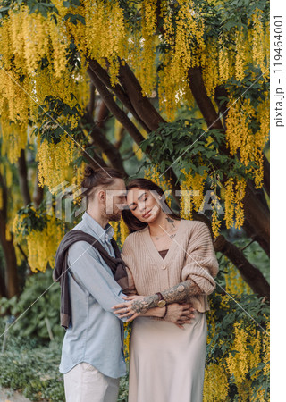 Amazing couple hugging under tree with yellow tree. Lovers couple hugging in a laburnum tree. Couple lovers hugging in the park. Girl with tattoos. A girl and a boy kiss. Amazing couple hugging under tree with yellow tree. Lovers couple hugging in a laburnum tree. Couple lovers hugging in the park. Girl with tattoos. A girl and a boy kiss. 119464001