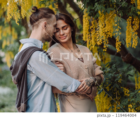 Amazing couple hugging under tree with yellow tree. Lovers couple hugging in a laburnum tree. Couple lovers hugging in the park. Girl with tattoos. A girl and a boy kiss. 119464004