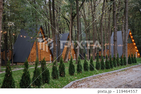 Cozy wooden A-frame cabins in forest with sunlight and outdoor seating area Cozy wooden A-frame cabins in forest with sunlight and outdoor seating area 119464557