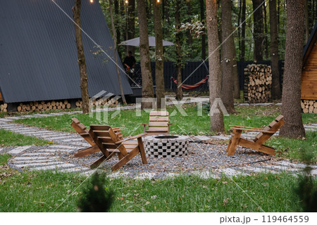 Cozy fire pit seating area in forest glamping with glowing string lights and modern wooden cabins 119464559