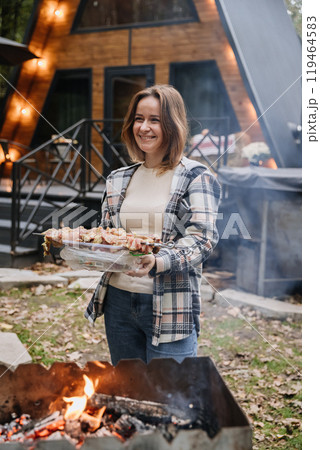 Woman preparing skewers for barbecue on an open fire, enjoying outdoor cooking with a smile 119464583