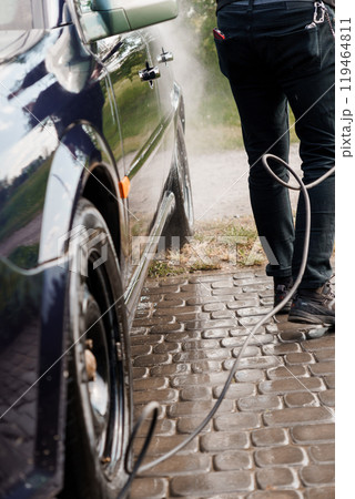 Person Using a Pressure Washer to Clean a Dark Blue Car on a Cobblestone Driveway 119464811