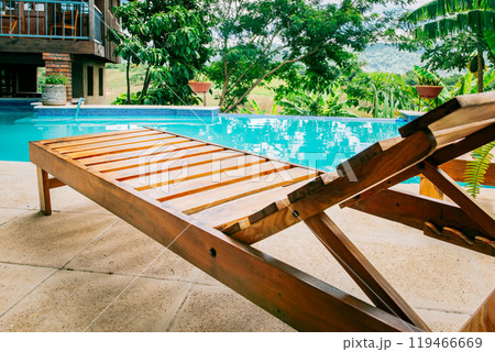 A wooden deck chair near a swimming pool. View of a Deck chair near a beautiful pool surrounded by nature 119466669