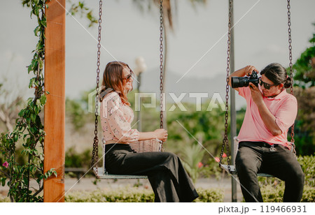 Boyfriend taking a photo of his girlfriend on a swing in a park. Boyfriend photographing girlfriend sitting on a swing 119466931