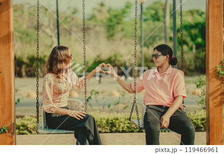Young couple in love sitting on a swing making a heart shape. Happy couple in love on a park swing making a heart shape Young couple in love sitting on a swing making a heart shape. Happy couple in love on a park swing making a heart shape 119466961