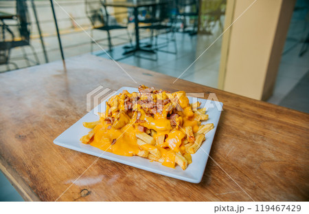 French fries dipped in yellow cheese with bacon on wooden table. Close up of french fries covered with cheddar cheese and bacon on wooden table 119467429