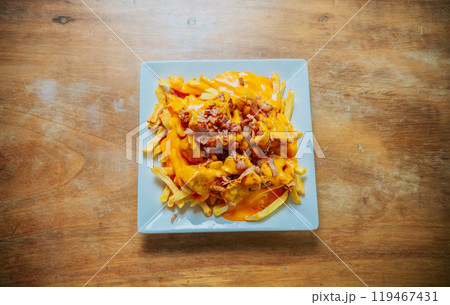 Top view of french fries covered with cheddar cheese and bacon on wooden table. French fries dipped in yellow cheese with bacon on wooden table 119467431