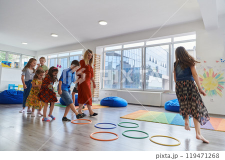 Small nursery school children with female teacher on floor indoors in classroom, doing exercise. Jumping over hula hoop circles track on the floor. 119471268