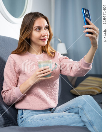 Young woman taking selfie while sitting on sofa with mug of tea Young woman taking selfie while sitting on sofa with mug of tea 119471404