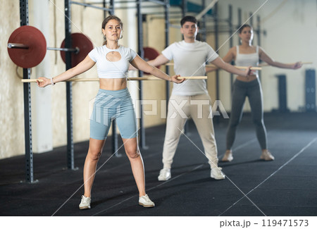 Slender girl, together with other athletes, does a warm-up using a long stick in gym Slender girl, together with other athletes, does a warm-up using a long stick in gym 119471573
