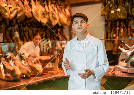 Young guy seller in white jacket selling spanish jamon in butcher shop Young guy seller in white jacket selling spanish jamon in butcher shop 119471732