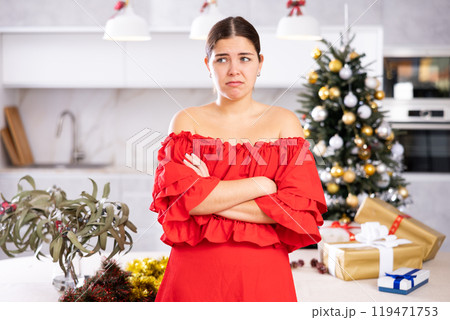 Portrait of sad young European woman in red dress posing in front of Christmas tree before holidays at flat Portrait of sad young European woman in red dress posing in front of Christmas tree before holidays at flat 119471753