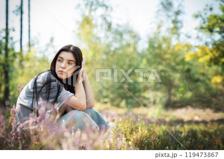 A young woman sitting thoughtfully in a serene forest clearing surrounded by colorful wildflowers  119473867