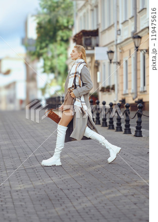 A woman walks confidently down a city street wearing stylish knee-high boots and a cozy jacket 119475616