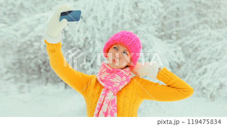 Happy cheerful smiling young woman taking selfie with smartphone in hat, sweater in snowy park 119475834