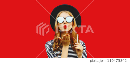 Portrait of beautiful young woman blowing her lips sends kiss with sweet red heart shaped lollipop Portrait of beautiful young woman blowing her lips sends kiss with sweet red heart shaped lollipop 119475842