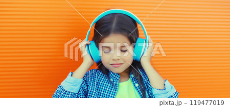 Happy little girl child in blue wireless headphones listening to music on orange background Happy little girl child in blue wireless headphones listening to music on orange background 119477019