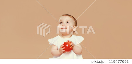 Portrait of cute baby holding red apple fruit sitting looking up on brown studio background 119477091
