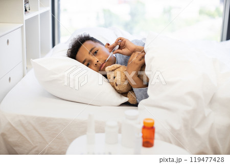African boy with flu lying in bed holding thermometer and teddy bear 119477428
