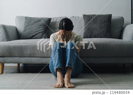 A woman is sitting on the floor in a living room. She is crying and she is in a state of distress A woman is sitting on the floor in a living room. She is crying and she is in a state of distress 119480034