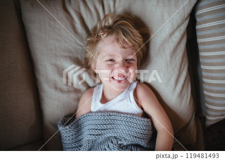 Sick boy with fever and cold lying on sofa, under blanket and smiling. 119481493