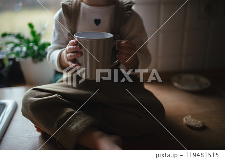 Adorable little girl is drinking tea. The sick girl with cold is sitting on kitchen counter with mug in hand. 119481515