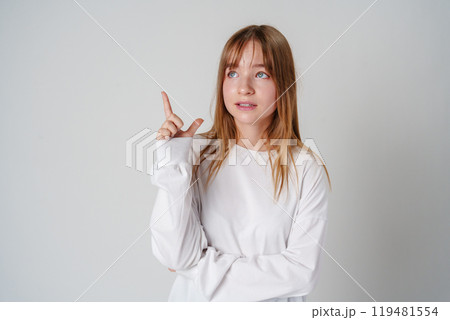 Young woman in a white long-sleeve shirt and blue jeans gestures thoughtfully indoors 119481554