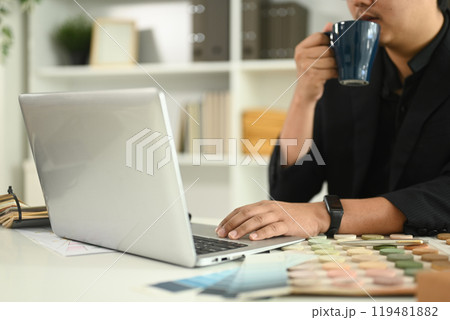 Male architect sipping coffee while working on a laptop at desk with model buildings and material samples Male architect sipping coffee while working on a laptop at desk with model buildings and material samples 119481882
