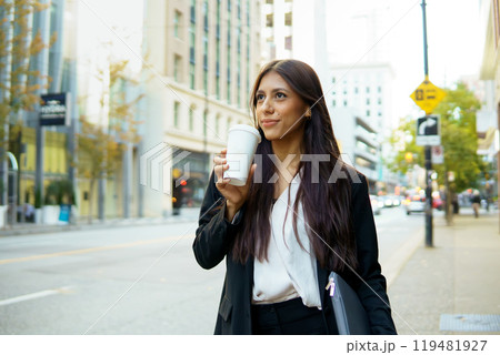 Smiling young businesswoman sipping from a takeaway coffee cup while walking in city street Smiling young businesswoman sipping from a takeaway coffee cup while walking in city street 119481927