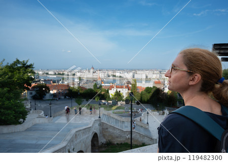 Budapest, Hungary. August 26, 2022. Aerial view of the city overlooking the Danube. A middle-aged woman gazes in fascination at the magnificent landscape. Travel destinations, travel lifestyle. Budapest, Hungary. August 26, 2022. Aerial view of the city overlooking the Danube. A middle-aged woman gazes in fascination at the magnificent landscape. Travel destinations, travel lifestyle. 119482300