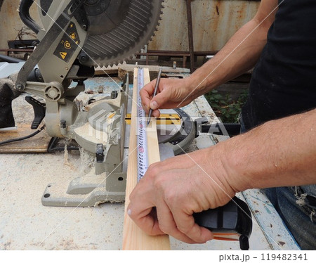male hands with tape measure and pencil measure length of wooden plank on workbench with miter saw, marking material before processing with circular saw male hands with tape measure and pencil measure length of wooden plank on workbench with miter saw, marking material before processing with circular saw 119482341